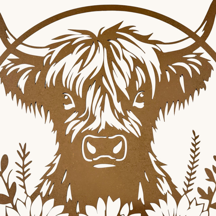 A close-up of a brown laser-cut metal wall art piece featuring a Highland cow head with long horns and decorative foliage, shown against a light off-white background.