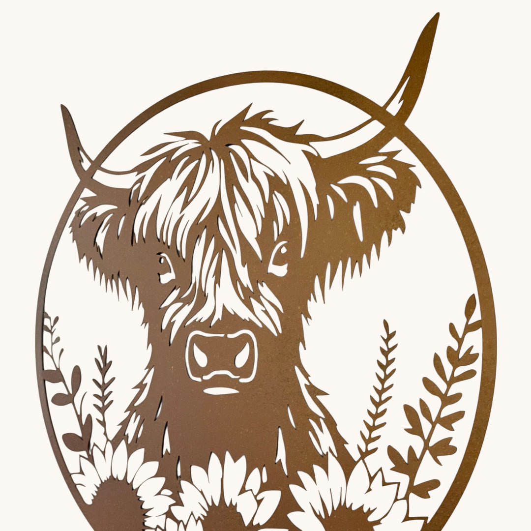 A circular metal wall art piece featuring a Highland cow face with sunflowers and leafy botanicals, shown mounted on a plain off-white wall.