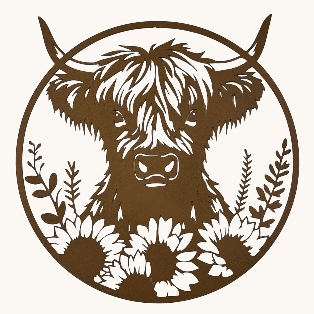 A circular metal wall art piece featuring a cutout Highland cow with flowers at the bottom, displayed against a plain off-white wall.