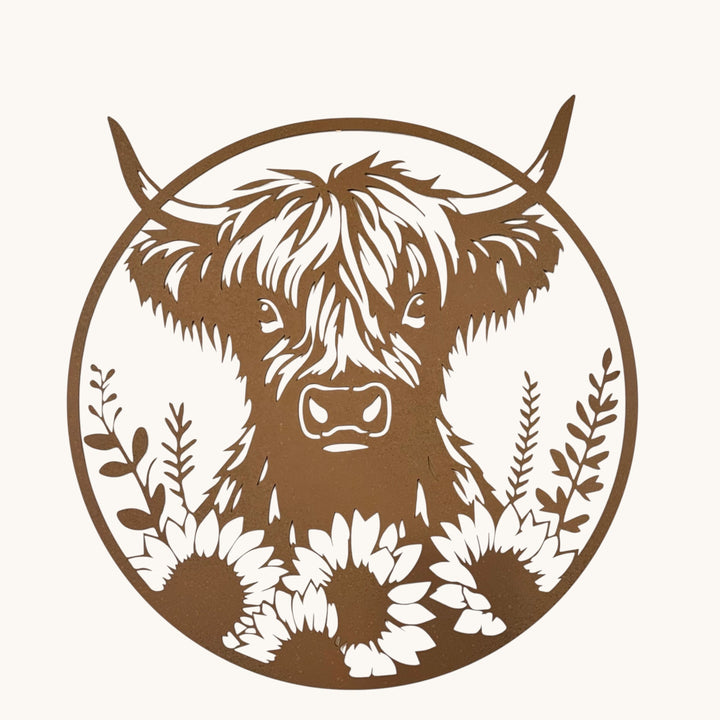 A circular metal wall art piece featuring a cutout Highland cow face with long horns and sunflower motifs at the bottom, displayed against a plain off-white wall.