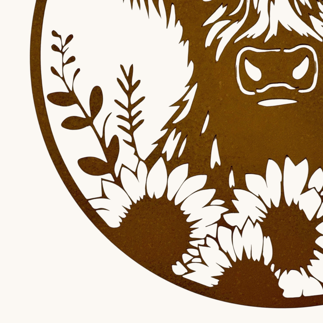 A close-up of a circular laser-cut metal wall art piece featuring a stylized face and sunflowers in a warm brown finish against a white textured background.