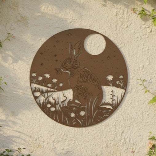 A circular metal garden wall art piece featuring a brown hare silhouette and a white moon, mounted on a textured cream stucco wall with green ivy around the edges.